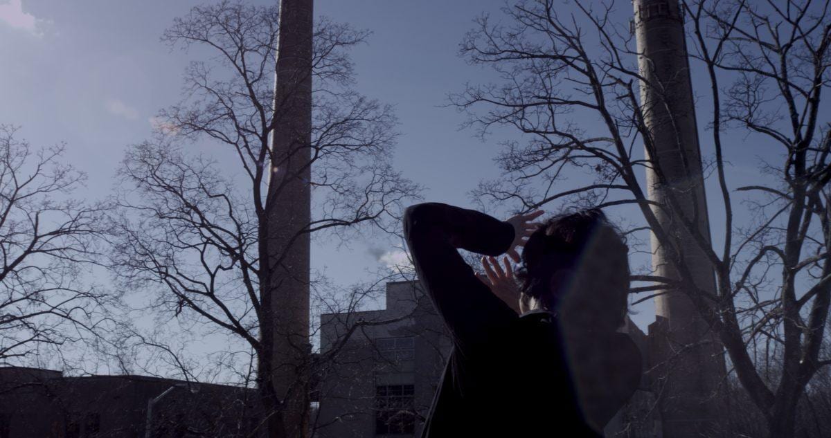 A woman sways toward an abandoned building against a stark sky and factory chimneys — Disturbance