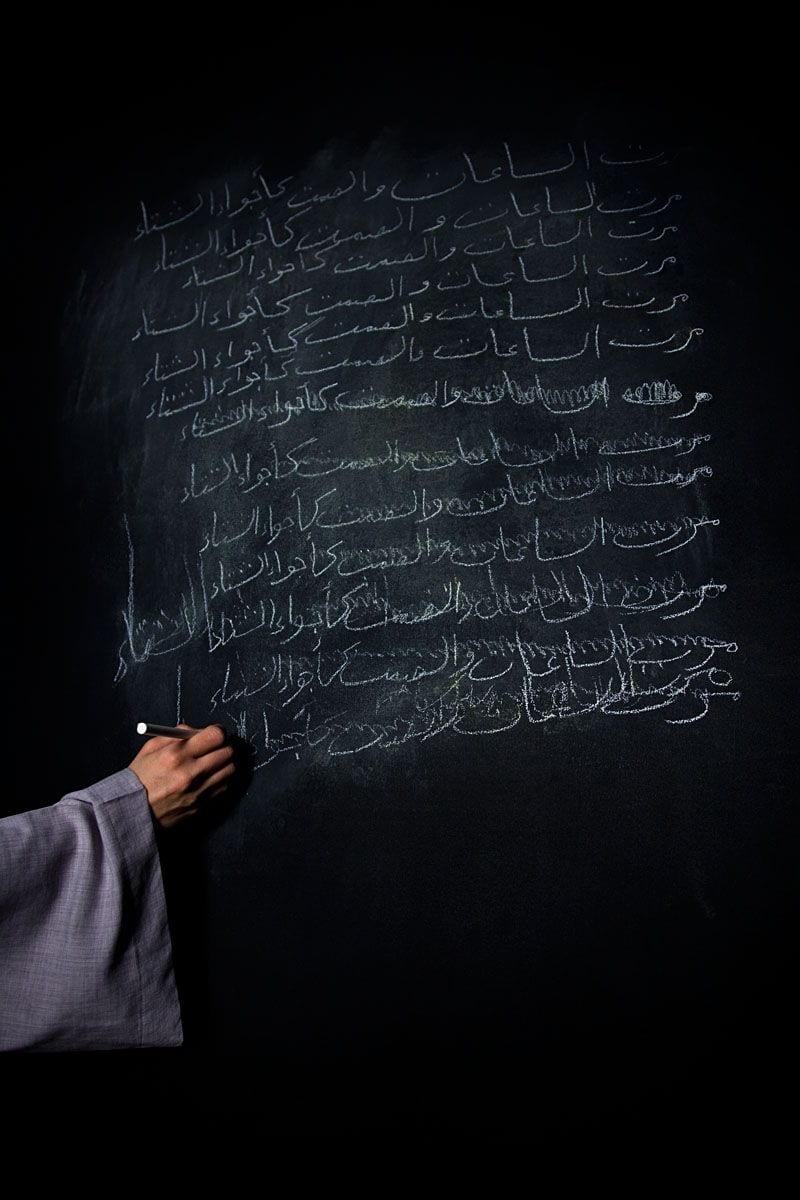 A hand writes a poem in chalk across a blackboard, the lines half-erased — Baghdadi Mem/Wars, Efface/Remain