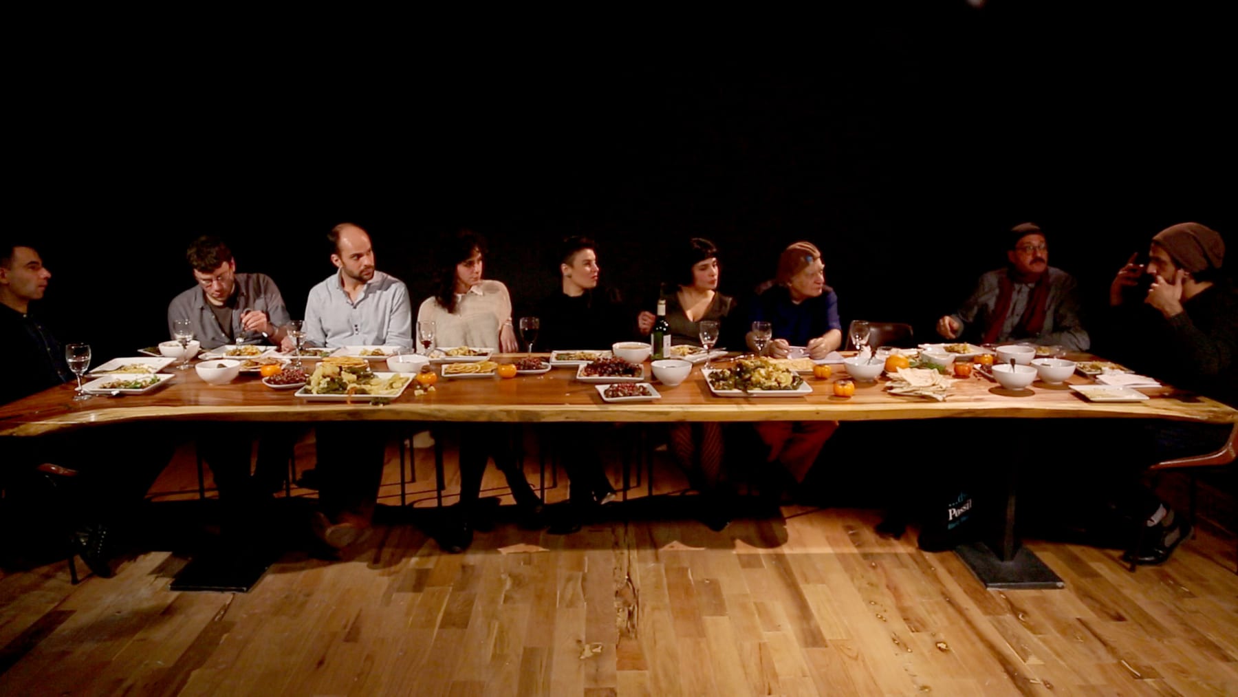 A long communal table of diasporic Iraqis sharing a meal in a darkened room — The Taste of Displacement