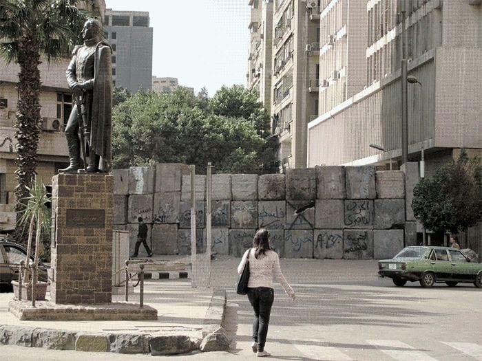 A figure walks past a colonial statue and concrete blast walls in central Cairo — This Stays Between Us